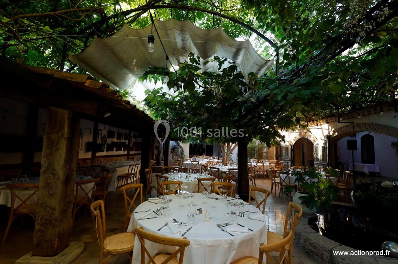 Salle de réception extérieure avec tables dressées sous une pergola ombragée par des arbres et une toile tendue.