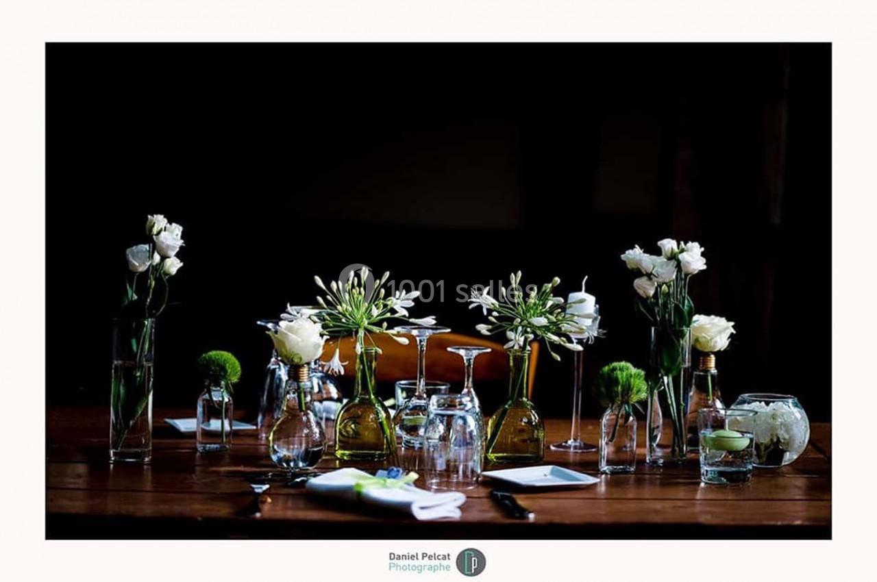 Table décorée avec des vases transparents contenant des fleurs blanches et vertes, entourée de verres et de bougies.