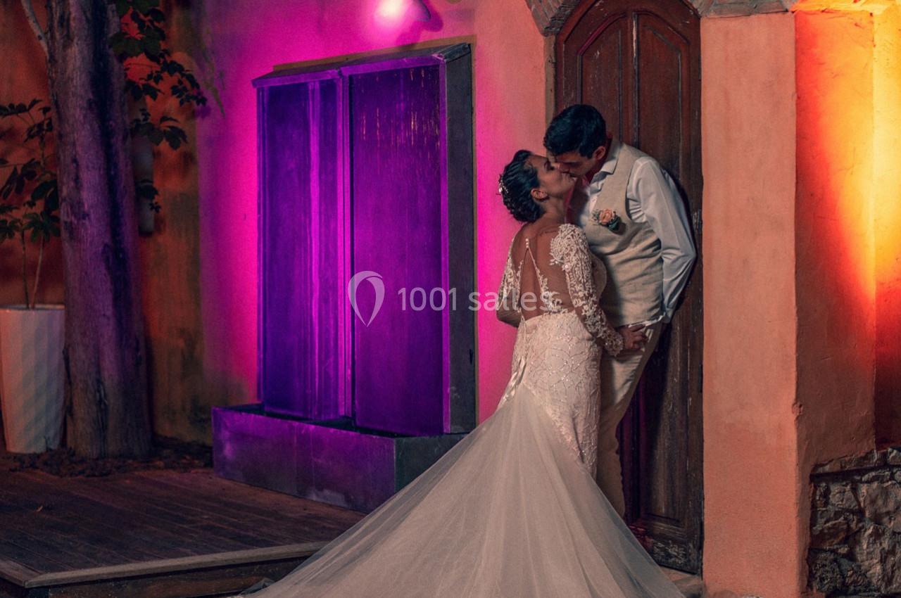 Un couple en tenue de mariage s'embrasse devant une porte en bois, éclairé par des lumières colorées.