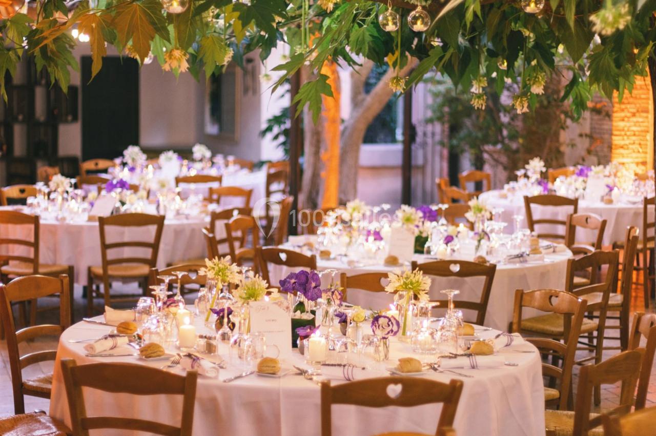 Tables rondes décorées de fleurs, bougies et vaisselle élégante dans un cadre extérieur avec feuillage suspendu.