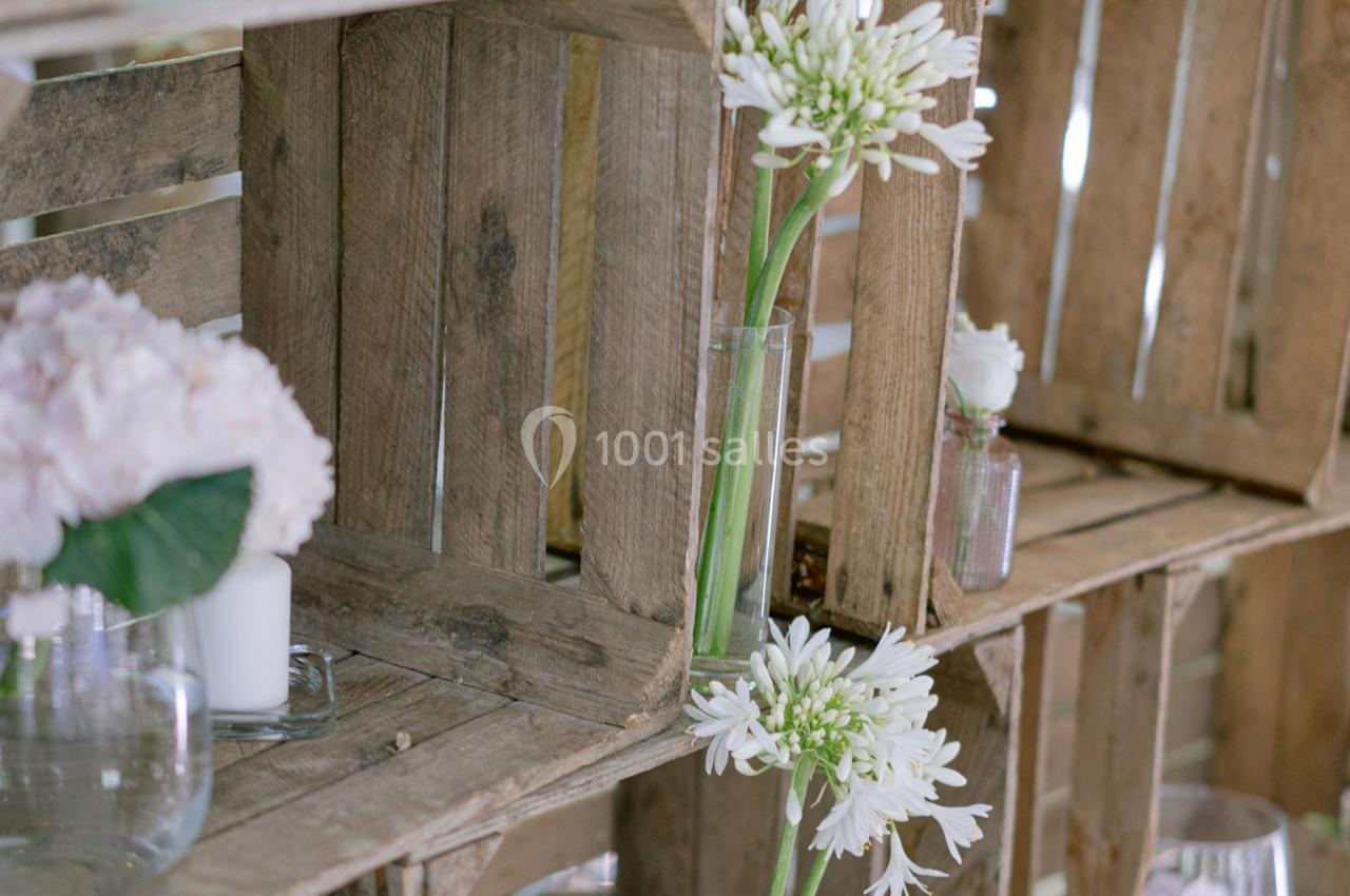 Des caisses en bois empilées contenant des vases avec des fleurs blanches et roses dans un décor rustique.