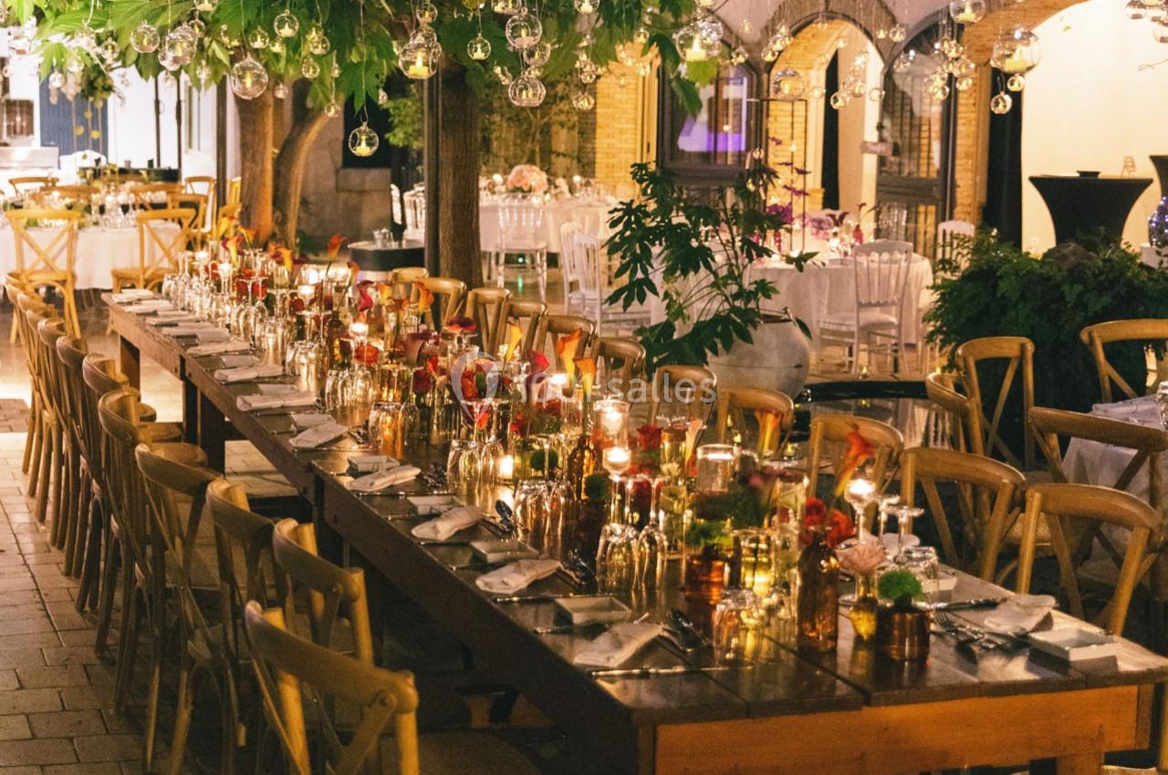 Grande table décorée pour un dîner en extérieur, entourée de chaises en bois, sous des lumières suspendues.