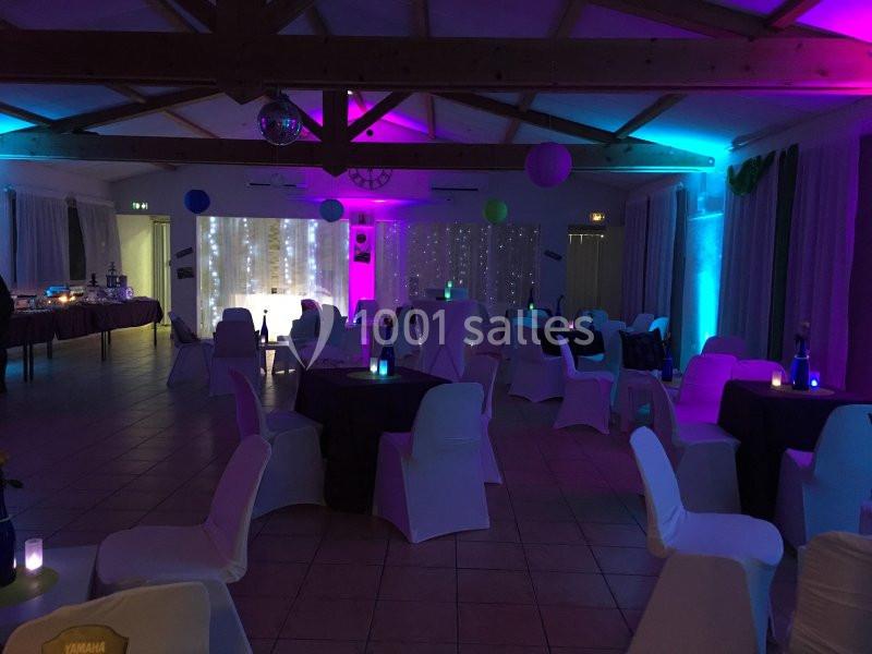 Salle de réception décorée avec des lumières colorées, des tables et des chaises blanches sous une charpente en bois.