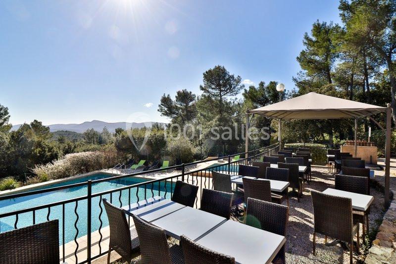 Terrasse extérieure avec tables et chaises en rotin, vue sur une piscine, des collines et des arbres sous un ciel ensoleillé.