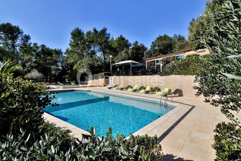 Piscine extérieure entourée de chaises longues, végétation et maison en arrière-plan sous un ciel dégagé.