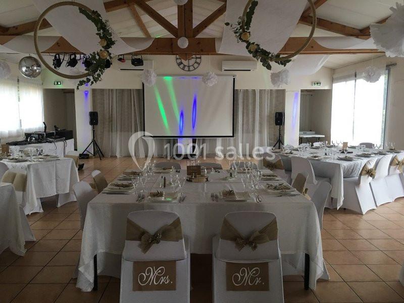 Salle de réception décorée pour un mariage, avec tables dressées, chaises ornées et écran de projection en fond.