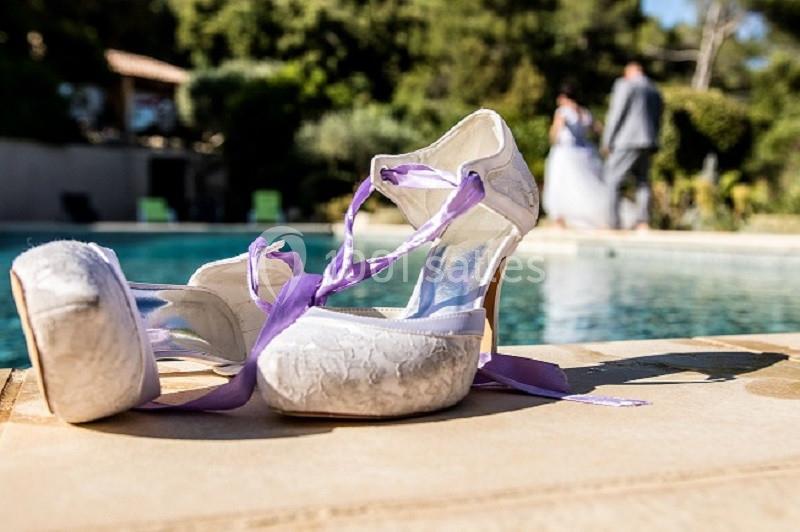 Chaussures de mariée blanches avec rubans violets posées près d'une piscine, en arrière-plan un couple flou.