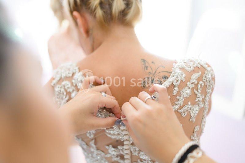 Une femme en robe de mariée avec un tatouage papillon dans le dos, tandis qu'une personne ajuste les boutons.