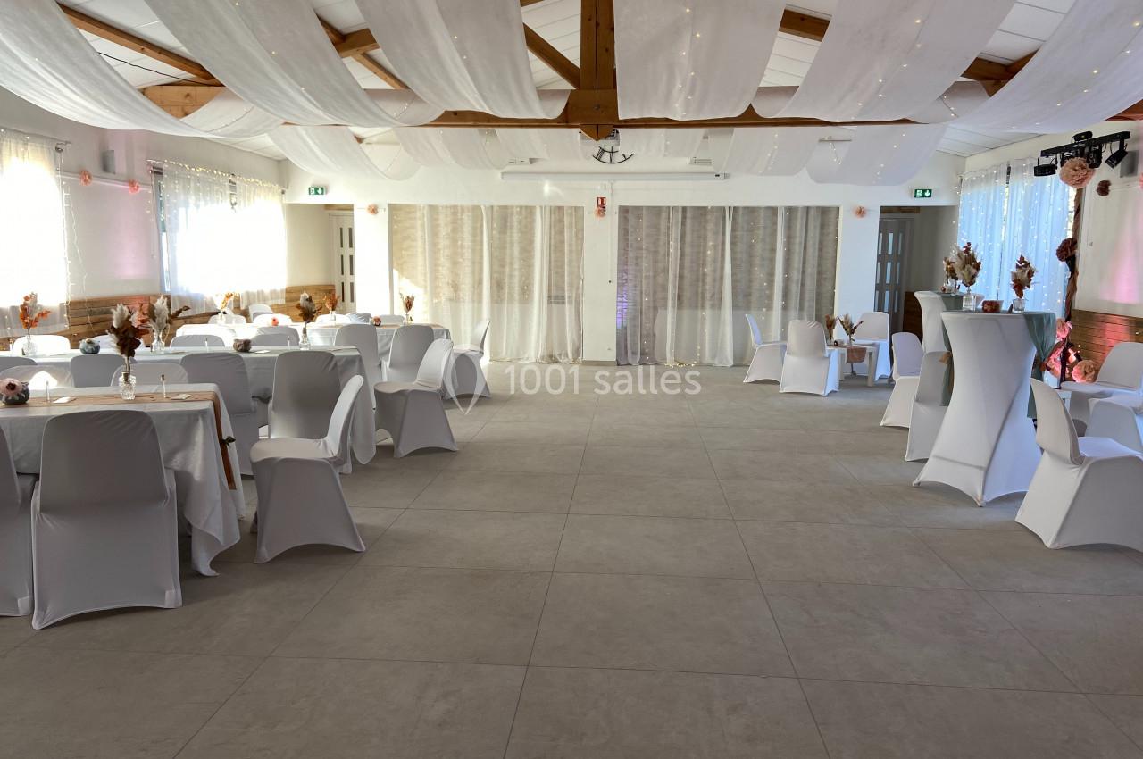 Salle de réception décorée avec des tables rondes, nappes blanches, chaises houssées et voilages suspendus au plafond.