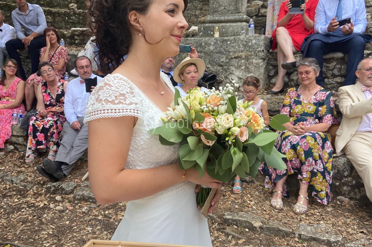 Une mariée en robe blanche tient un bouquet de fleurs devant des invités assis en plein air.