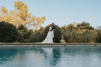 Un couple en tenue de mariage coupe une pièce montée décorée, devant un mur en pierre.