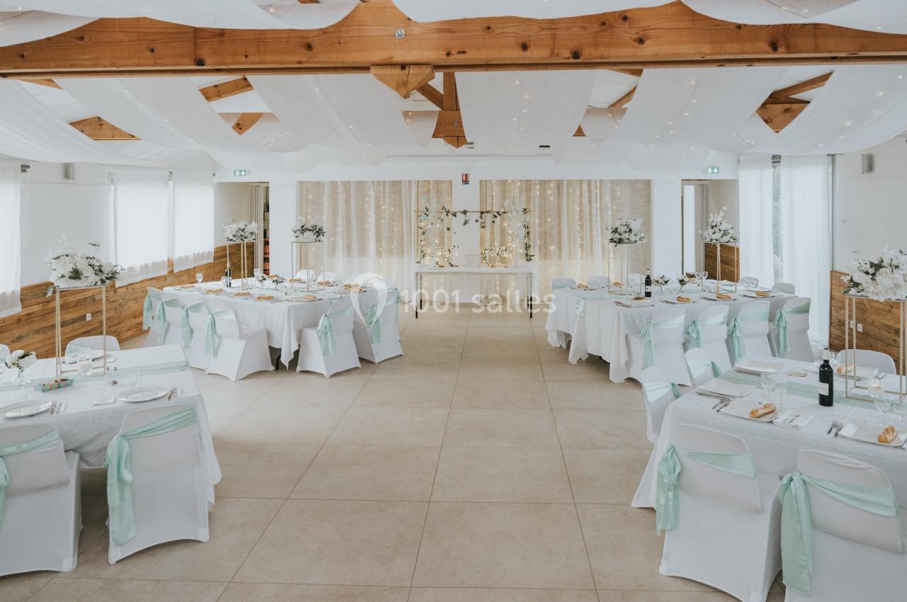 Salle de réception décorée avec des tables dressées, des chaises blanches ornées de rubans verts et un plafond drapé.