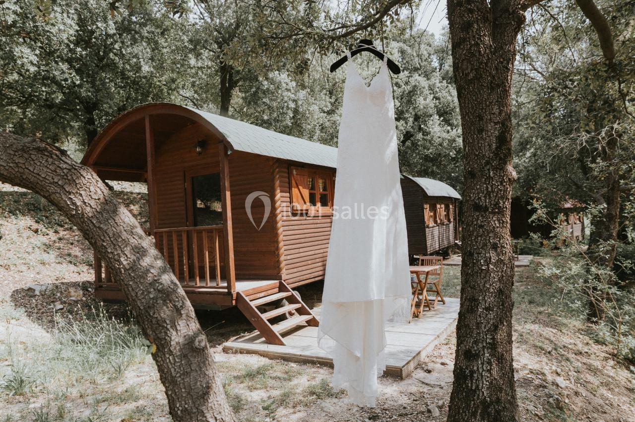 Robe de mariée blanche suspendue à une branche devant des roulottes en bois dans un cadre boisé.