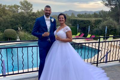Un couple en tenue de mariage coupe une pièce montée décorée, devant un mur en pierre.