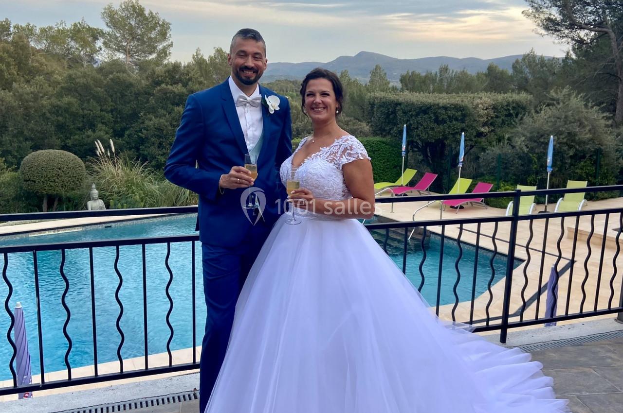 Un couple en tenue de mariage pose devant une piscine extérieure avec un paysage arboré en arrière-plan.