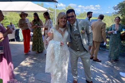 Un couple en tenue de mariage coupe une pièce montée décorée, devant un mur en pierre.