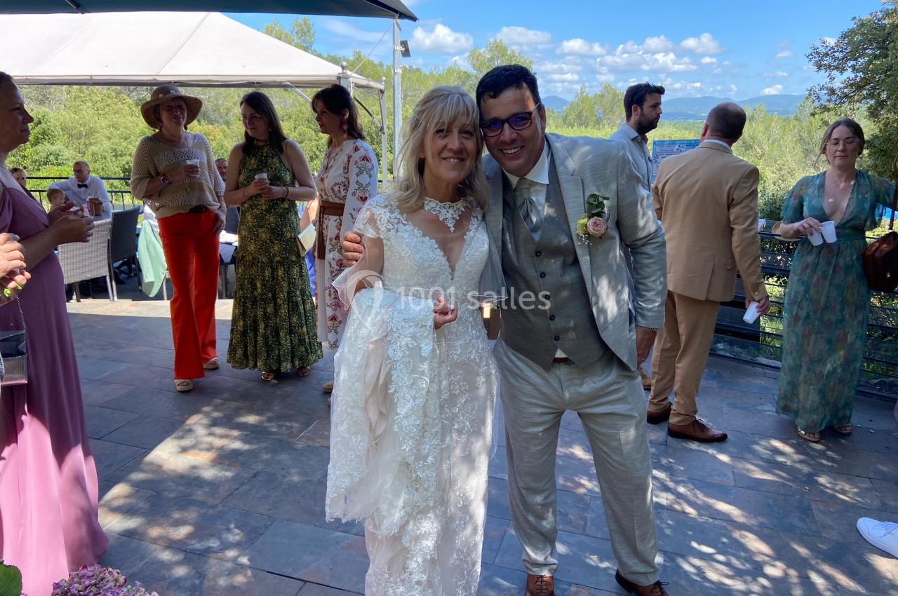 Un couple en tenue de mariage sourit sous un ciel bleu, entouré d'invités sur une terrasse extérieure.