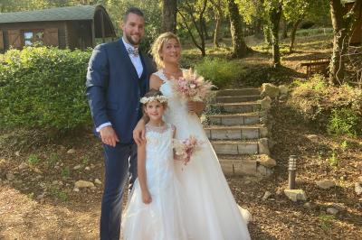 Un couple en tenue de mariage coupe une pièce montée décorée, devant un mur en pierre.