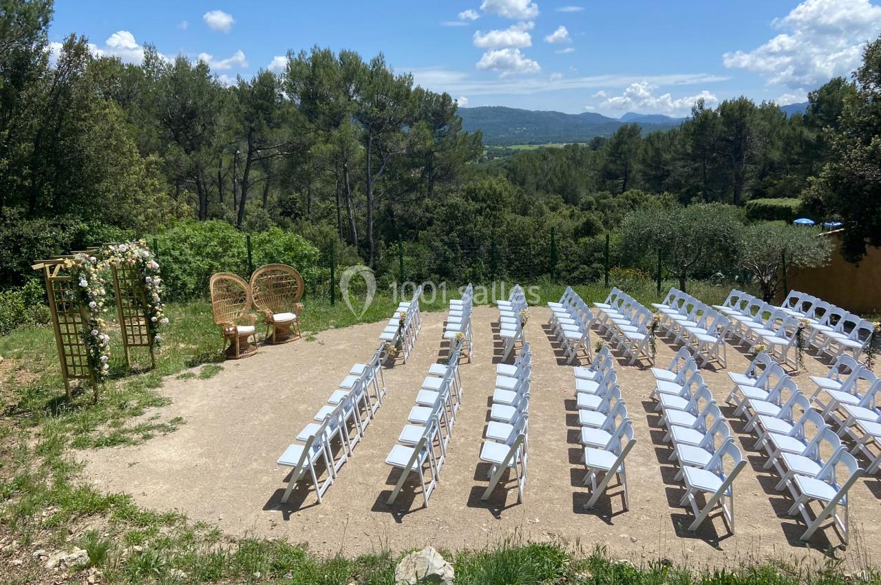 Chaises blanches disposées en rangées face à une arche fleurie dans un cadre naturel avec vue sur des collines boisées.