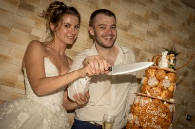 Un couple en tenue de mariage coupe une pièce montée décorée, devant un mur en pierre.