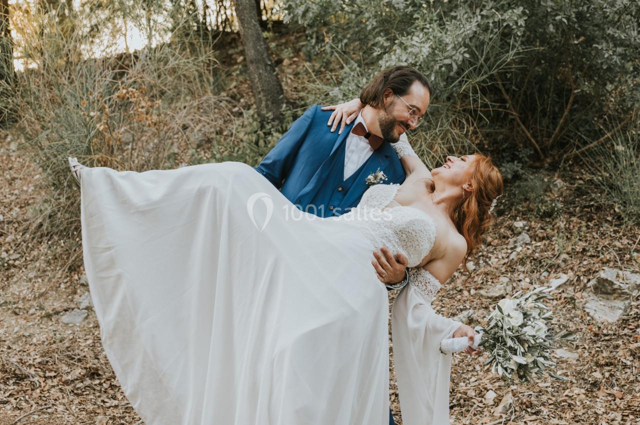 Un homme en costume bleu porte une femme en robe de mariée dans un cadre naturel boisé.