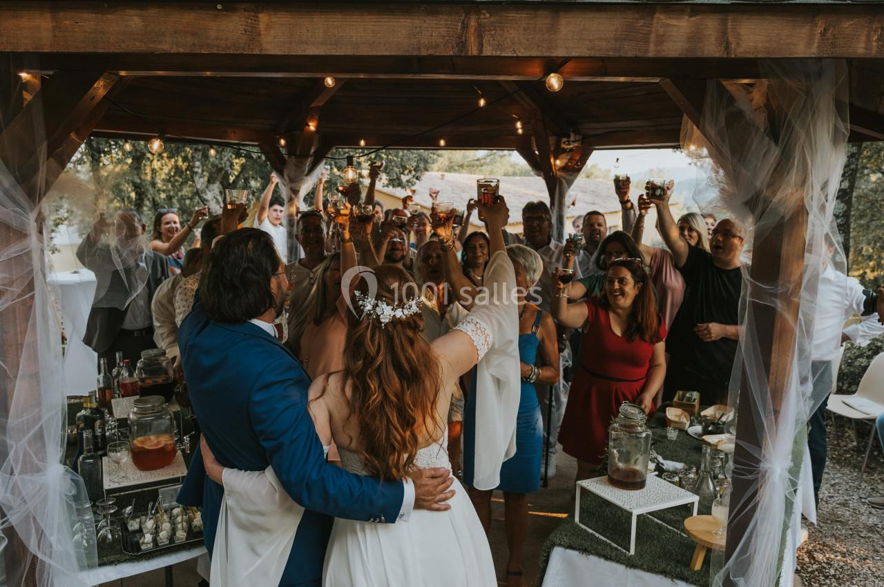Un couple de mariés lève un verre avec des invités sous une pergola décorée, lors d'une célébration en plein air.