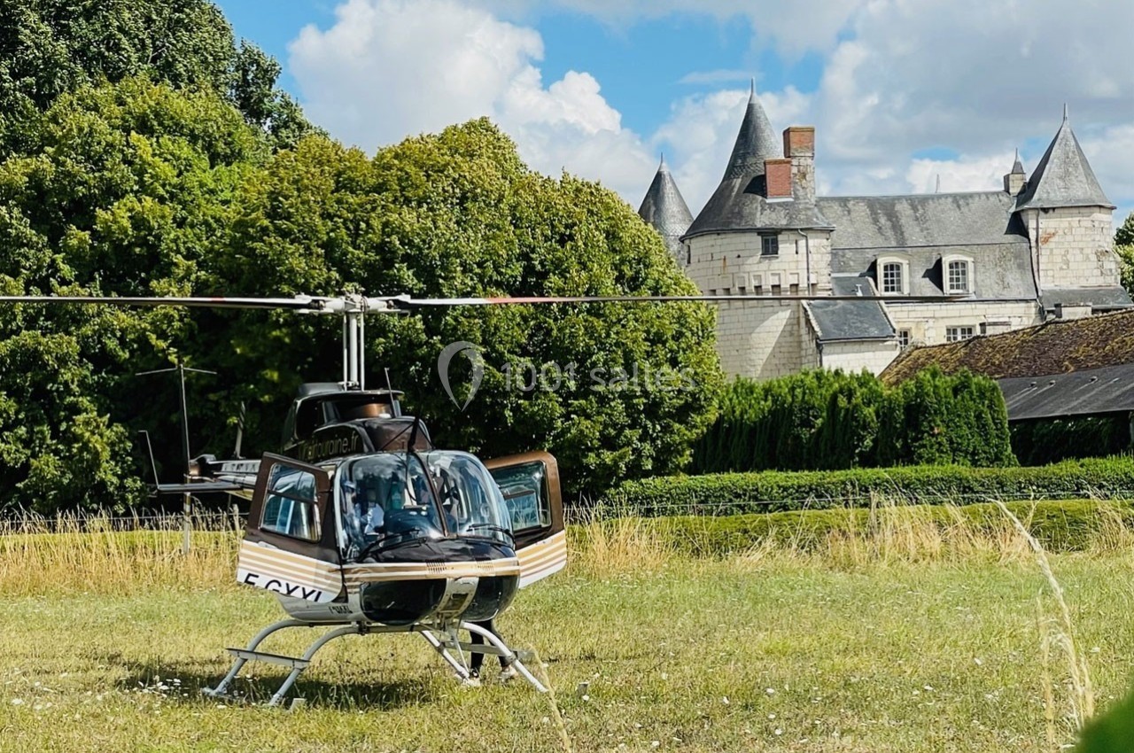 Hélicoptère posé dans un champ verdoyant avec un château en arrière-plan sous un ciel partiellement nuageux.