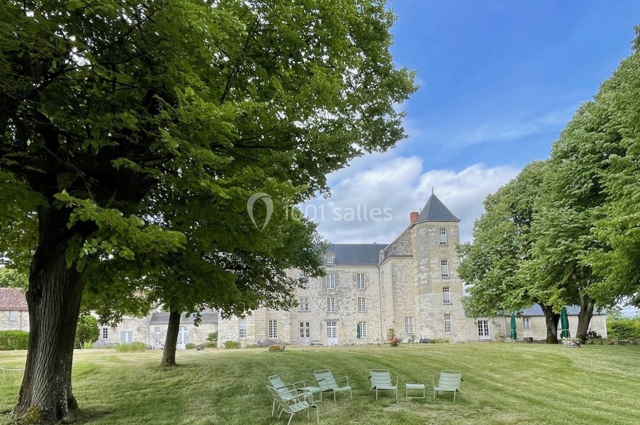 Château en pierre entouré d'arbres, avec des chaises disposées sur une pelouse sous un ciel partiellement nuageux.