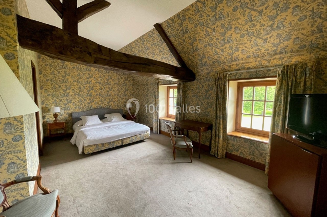 Chambre lumineuse avec lit double, poutres apparentes, murs tapissés, bureau, fauteuil et fenêtres donnant sur un jardin.