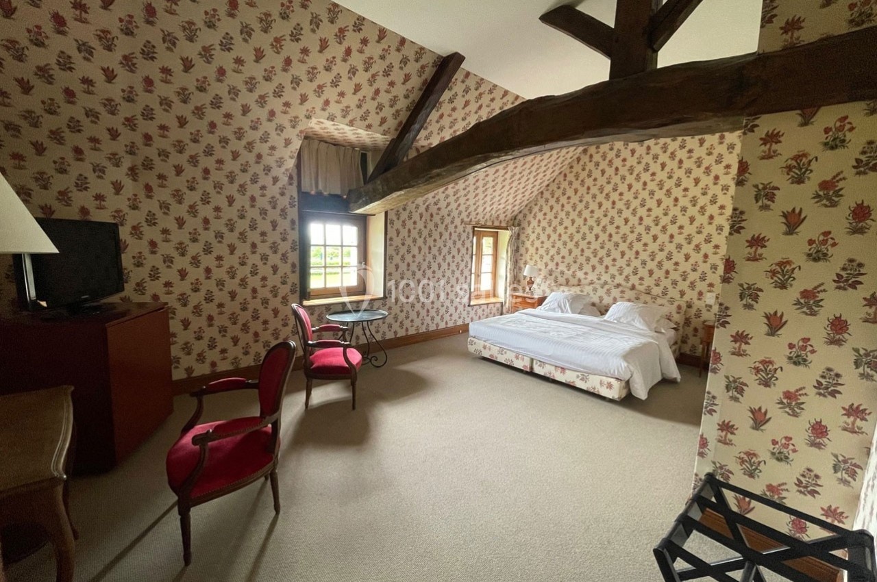 Chambre mansardée avec poutres apparentes, lit double, mobilier classique et murs décorés de papier peint fleuri.
