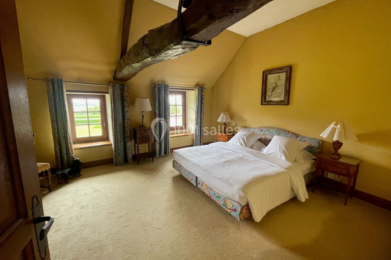 Chambre lumineuse avec lit double, poutre apparente, murs jaunes, deux fenêtres et mobilier en bois.