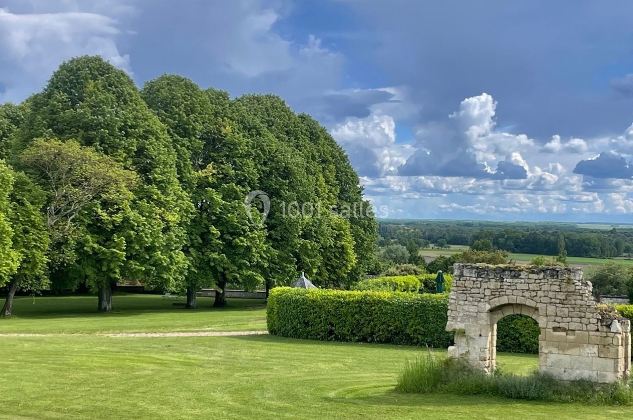 Ruine en pierre dans un parc verdoyant avec pelouse, arbres et ciel nuageux en arrière-plan.