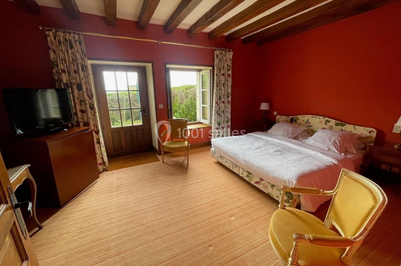 Chambre lumineuse avec murs rouges, lit double, fauteuils jaunes, télévision et fenêtres donnant sur un jardin.