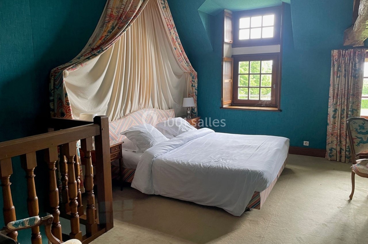 Lit double avec baldaquin, draps blancs et rideaux colorés dans une chambre aux murs bleus avec fenêtre et escalier en bois.