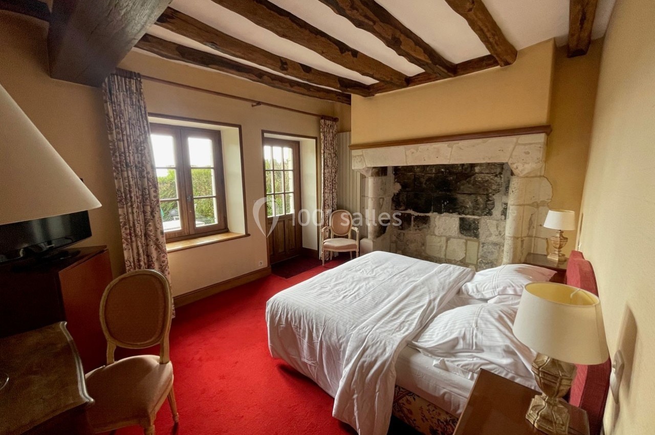 Chambre avec lit double, poutres apparentes, cheminée en pierre, moquette rouge et vue sur un jardin par une fenêtre.