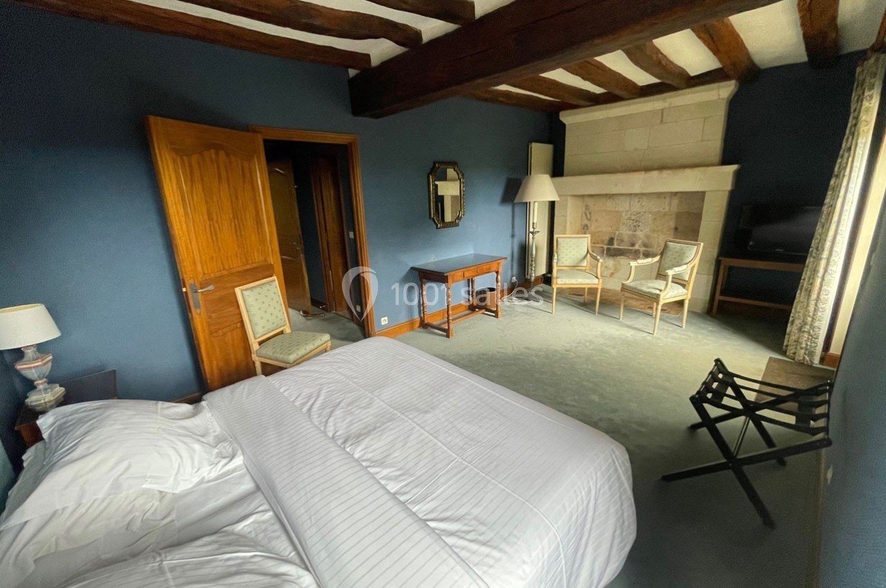 Chambre avec lit double, murs bleus, poutres apparentes, cheminée en pierre, et mobilier classique.