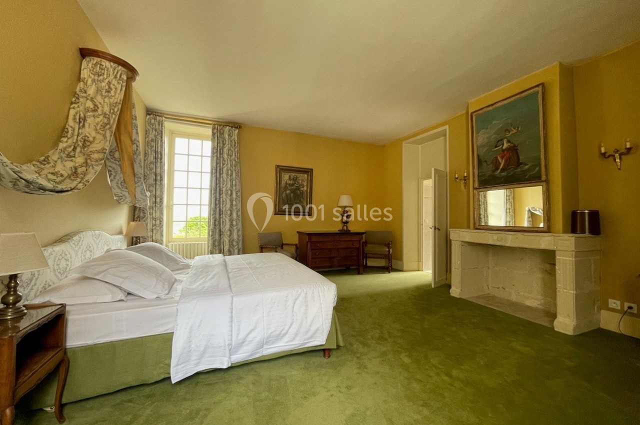 Chambre lumineuse avec lit double, cheminée en pierre, murs jaunes, moquette verte et grande fenêtre donnant sur l'extérieur.