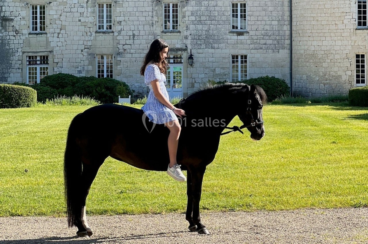 Une femme en robe bleue est assise sur un cheval noir immobile devant un bâtiment en pierre.