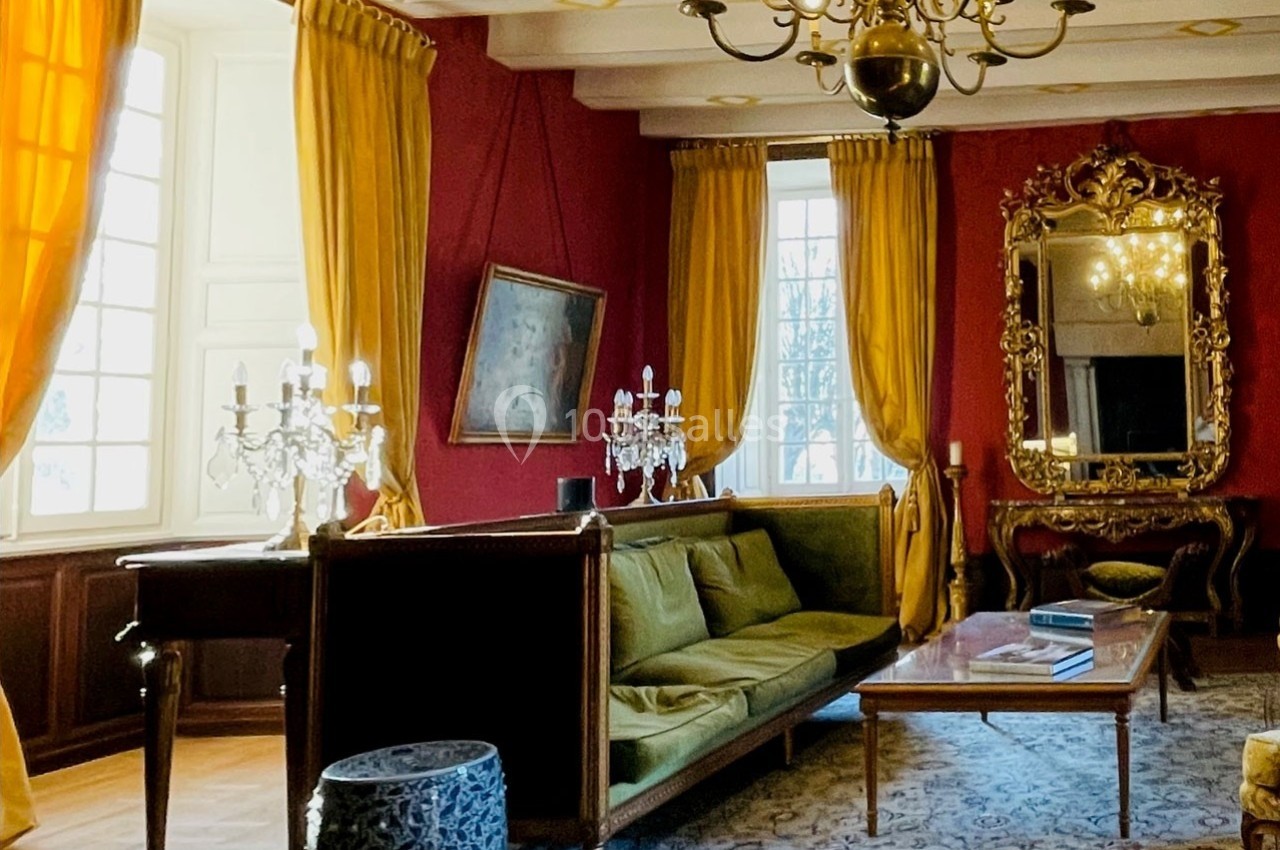 Salon élégant avec murs rouges, rideaux dorés, canapé vert, lustres, miroir doré et lumière naturelle.