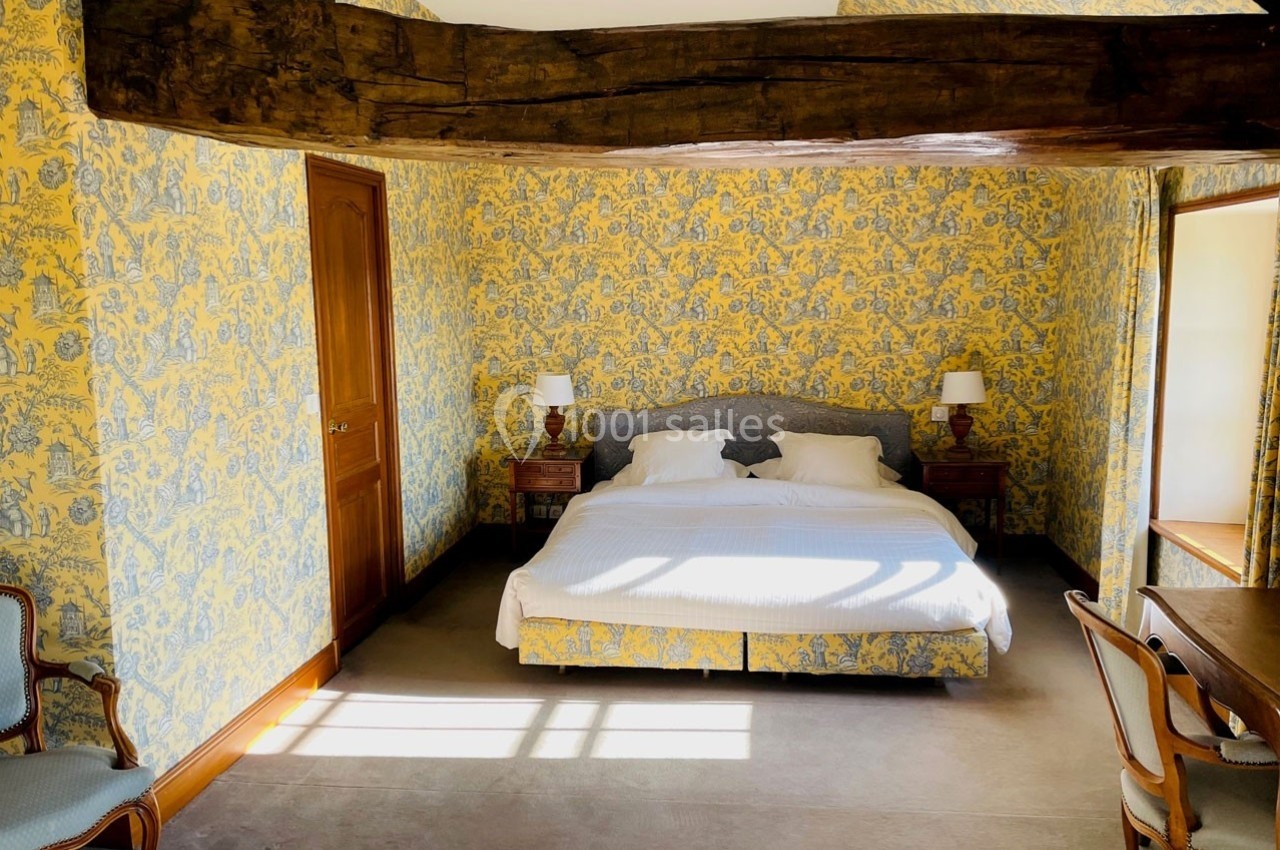 Chambre lumineuse avec lit double, poutre apparente, murs recouverts de papier peint jaune à motifs et mobilier en bois.