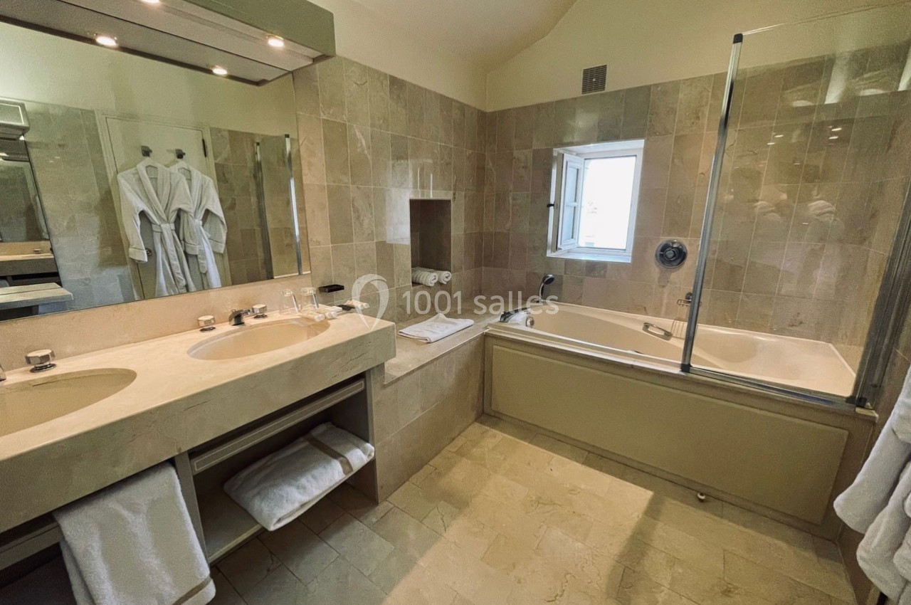 Salle de bain avec baignoire, double vasque, miroir éclairé, peignoirs accrochés et carrelage beige.