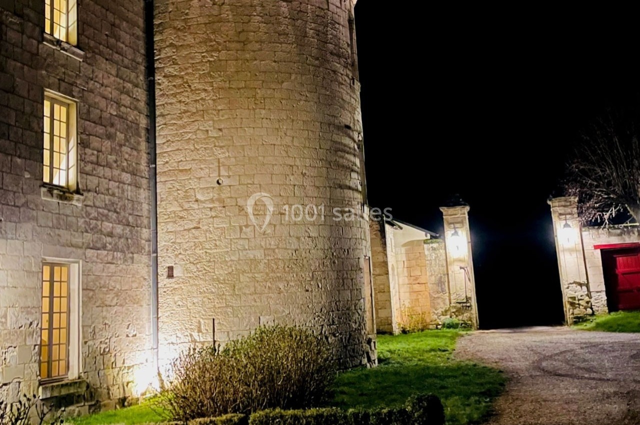 Façade éclairée d'un bâtiment en pierre avec une tour ronde, une allée et un portail ouvert de nuit.