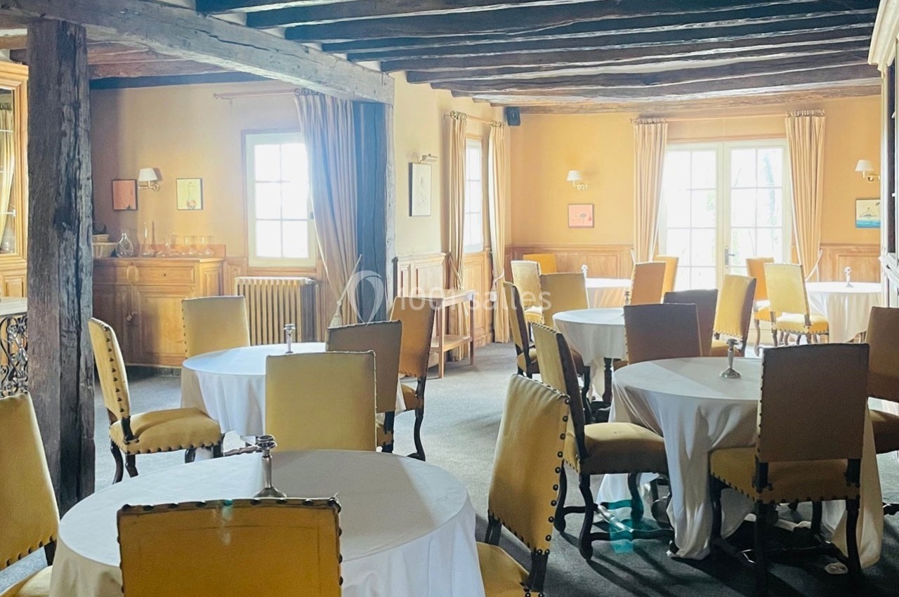 Salle de restaurant lumineuse avec tables rondes dressées, chaises rembourrées et poutres apparentes au plafond.