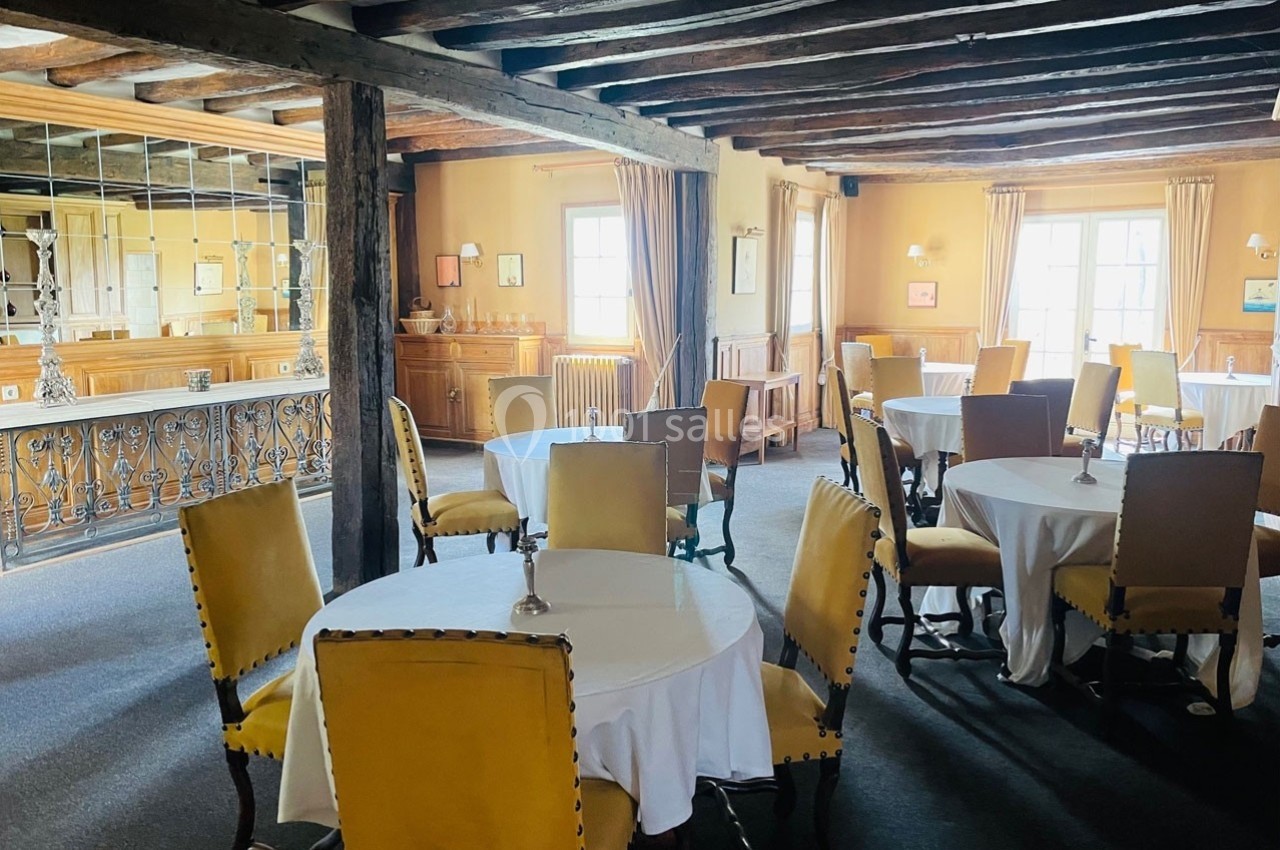 Salle à manger lumineuse avec des tables rondes dressées, des chaises jaunes et un plafond en poutres apparentes.