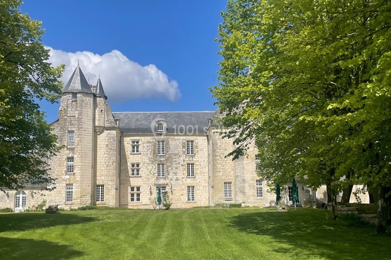 Façade d'un château en pierre avec une tour, entouré d'arbres et d'une pelouse sous un ciel bleu.