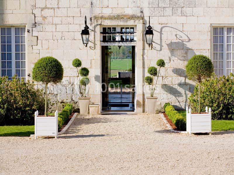 Entrée d'un bâtiment en pierre avec porte ouverte, lanternes murales et jardinières de buis taillés.