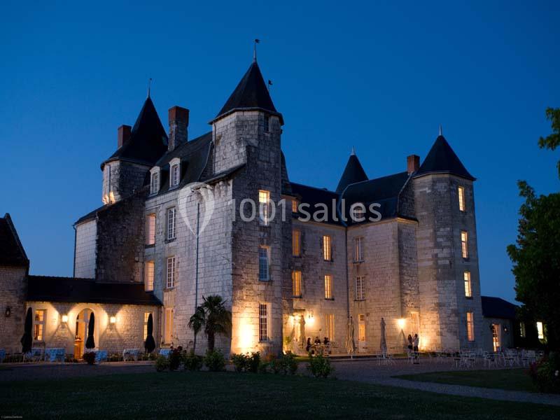 Château en pierre éclairé à la tombée de la nuit, avec des tours et des fenêtres illuminées.