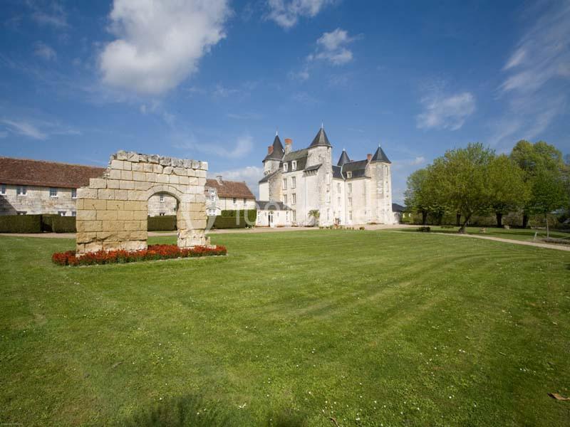 Château en pierre avec tours, pelouse verdoyante, ruines d'un mur en premier plan et ciel bleu dégagé.