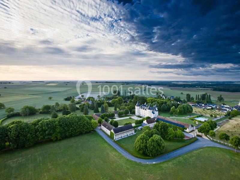 Vue aérienne d'un château entouré de jardins verdoyants et de champs sous un ciel partiellement nuageux.