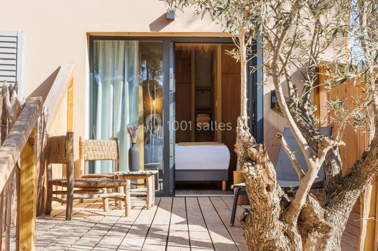 Terrasse en bois avec chaises en osier, table basse et vue sur une chambre à travers une baie vitrée ouverte.